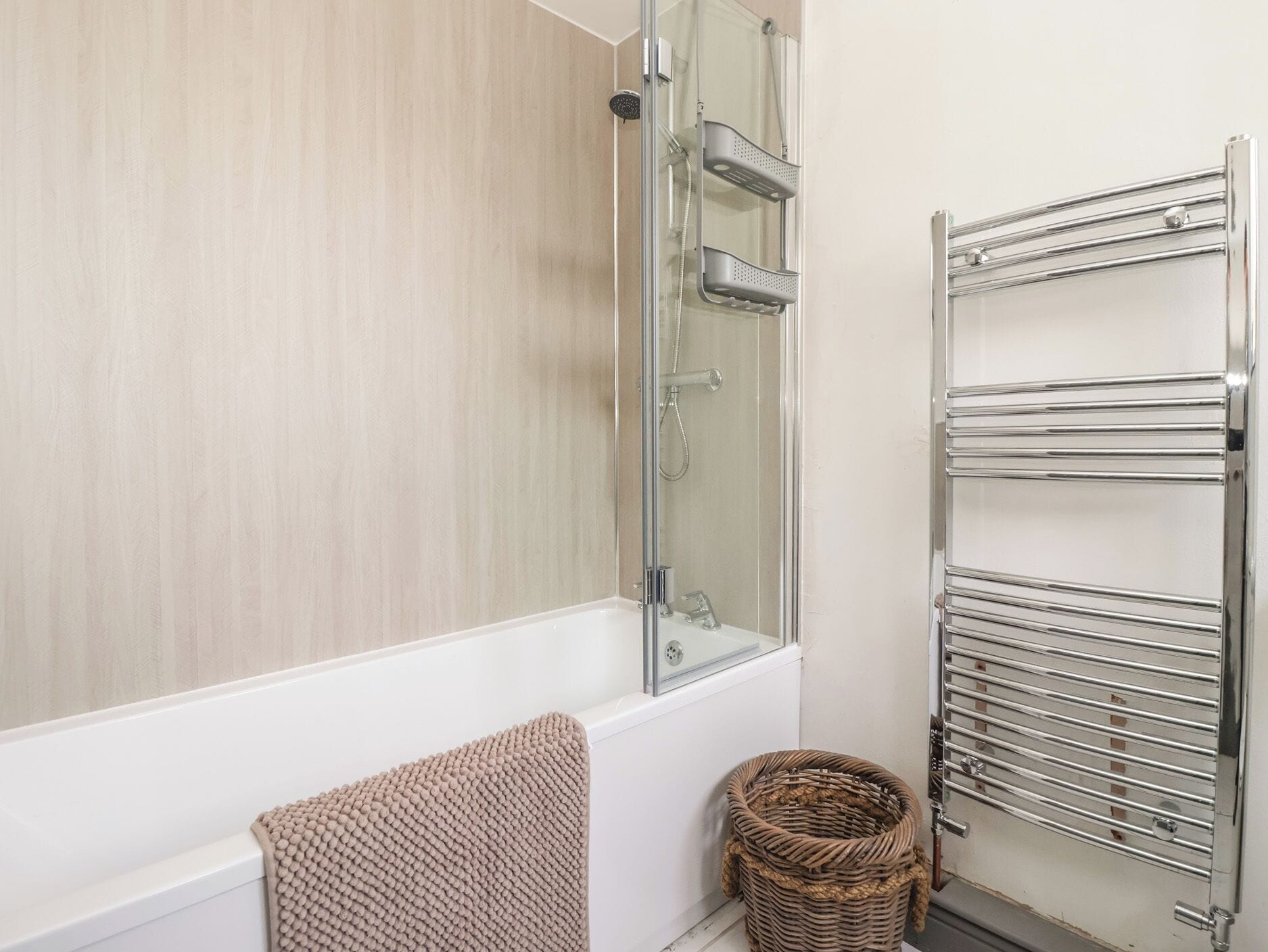 A bathroom with a glass-enclosed shower, a white bathtub, a chrome towel rack, and a wicker basket.