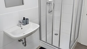Kylpyhuoneessa on lasiseinällä varustettu suihku, valkoinen pesuallas, jonka yläpuolella on peili, sekä musta matto lattialla.