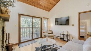 A spacious living room with a large sliding glass door to a deck, a flat-screen TV mounted on the wall, a round glass coffee table, and a cozy sofa.