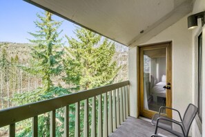 A balcony with a wooden railing, a door leading to a room with a bed, and a view of trees and hills.