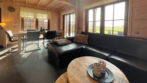 A spacious room with a wooden ceiling, a black leather sofa, a dining table with chairs, and large windows offering a view of the outdoors. - Forest Villa, Rural Twente (Den Ham)