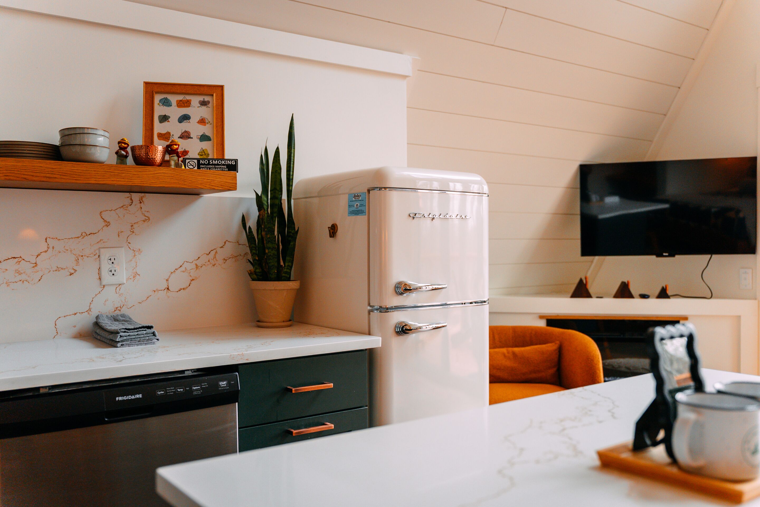 A kitchen with a white refrigerator, a dishwasher, a marble countertop, a plant, and a television mounted on the wall.