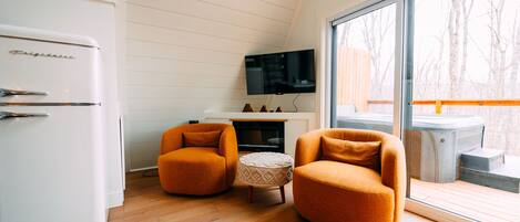 A modern kitchen with a white refrigerator, two orange armchairs, a small round ottoman, and a flat-screen TV mounted on the wall.