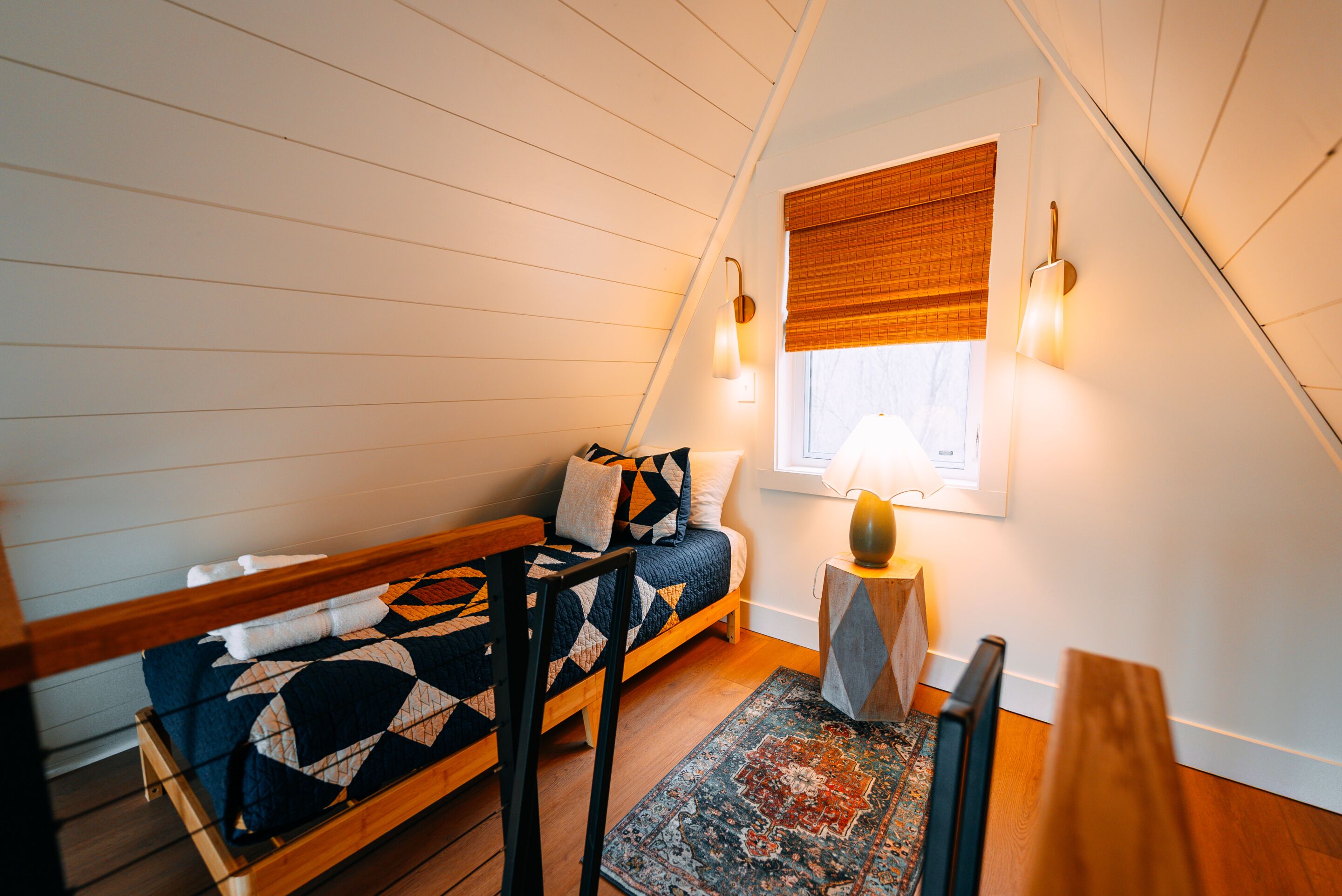 A loft bedroom with a wooden bed, a window with blinds, a nightstand with a lamp, and a patterned rug.