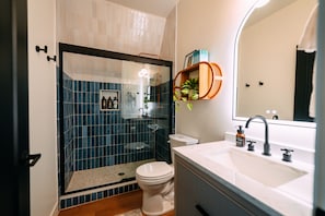 A modern bathroom with a glass-enclosed shower, a white toilet, a sink with a mirror, and a shelf with toiletries. - Hickory Hollow | Hocking Hills, Hot Tub, Romantic (McArthur)