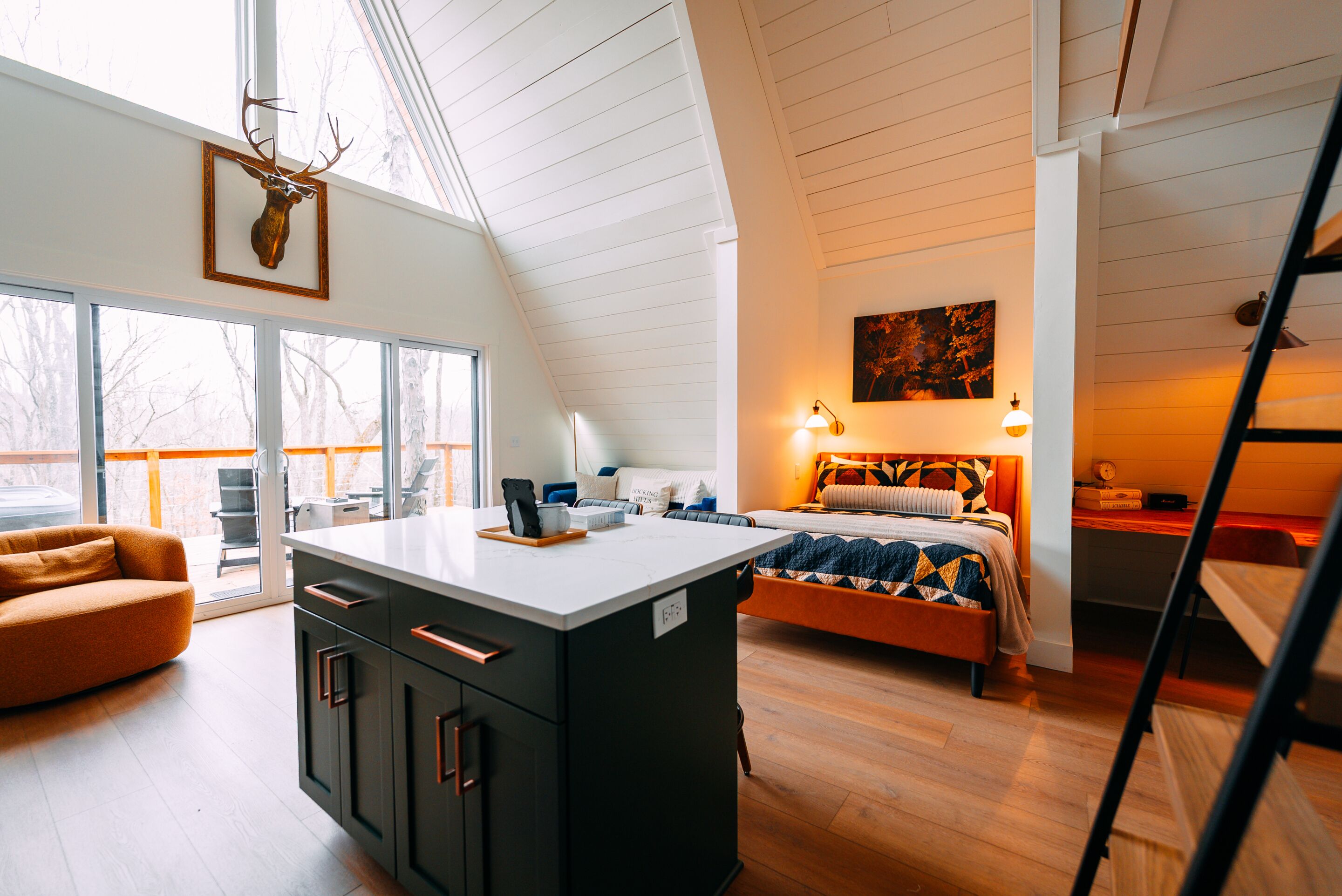A modern interior with a loft-style ceiling, a central island with cabinets, a bed with a patterned bedspread, and a painting on the wall.