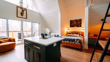 A modern interior with a loft-style ceiling, a central island with cabinets, a bed with a patterned bedspread, and a painting on the wall.