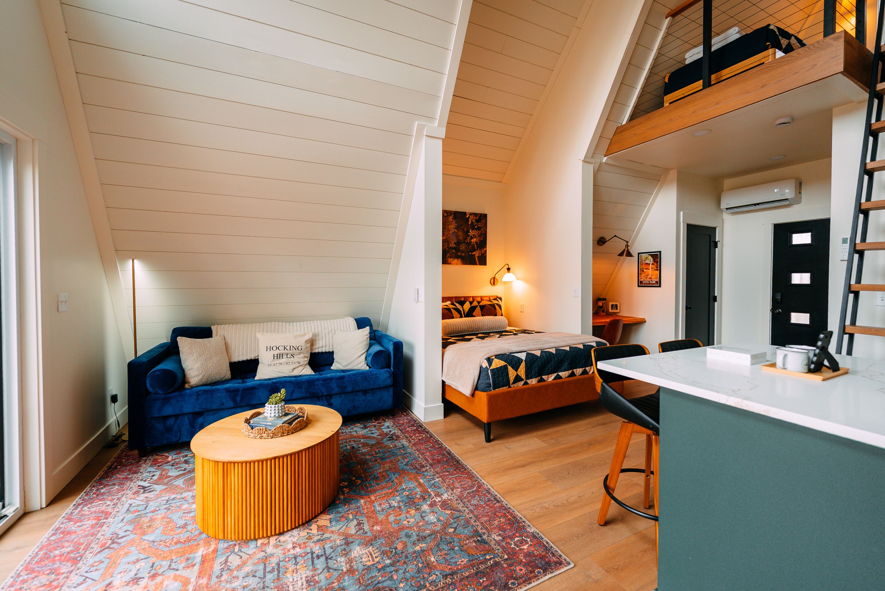 A loft-style room with a sloped ceiling, a blue sofa, a wooden coffee table, a bed with patterned bedding, and a small kitchenette area.