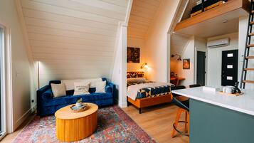 A loft-style room with a sloped ceiling, a blue sofa, a wooden coffee table, a bed with patterned bedding, and a small kitchenette area.