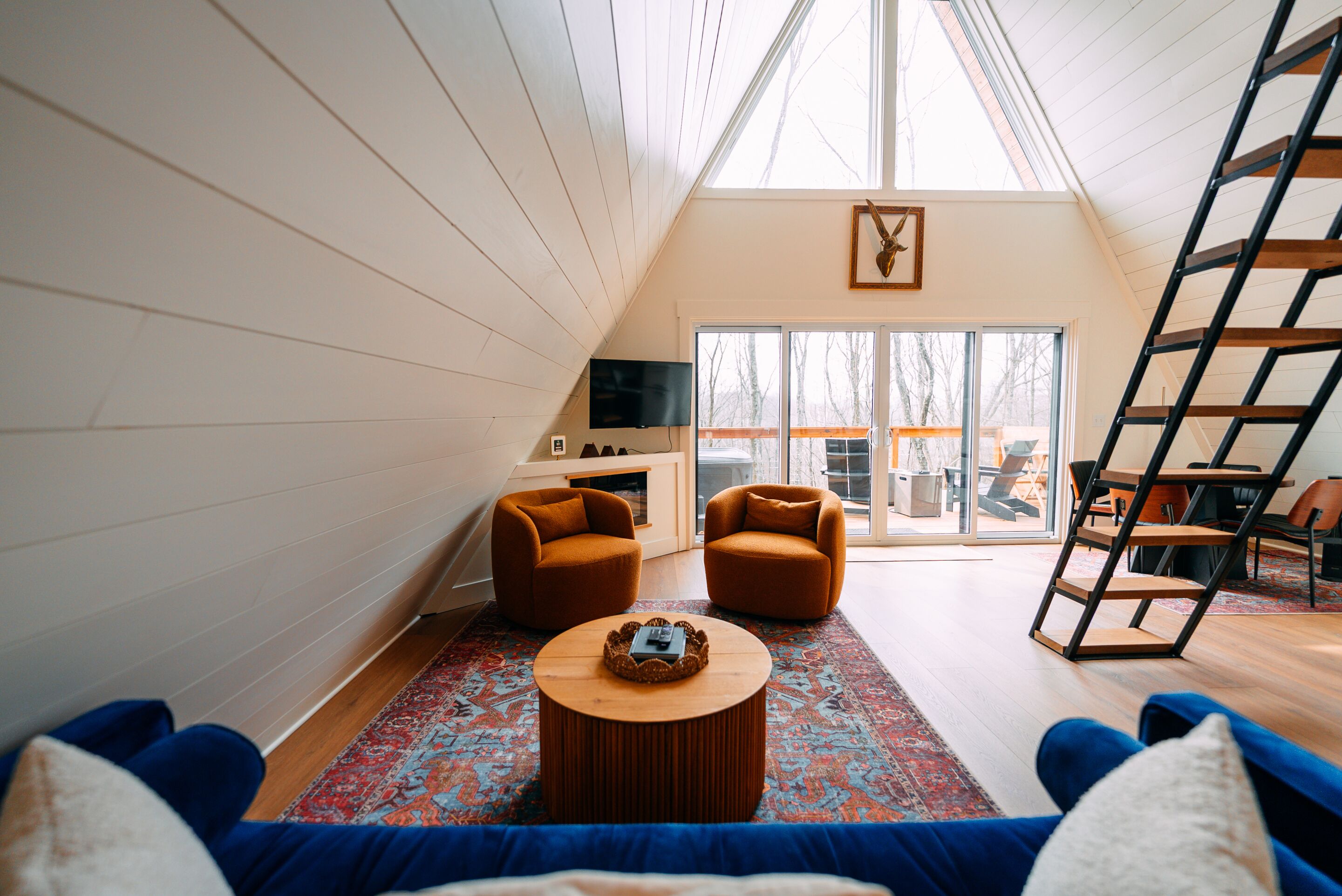A loft-style room with a high ceiling, a large window, a TV, two sofas, a coffee table, and a ladder.