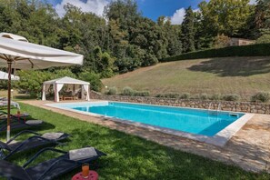 Piscina con sillas de descanso y una carpa, rodeada de un muro de piedra y vegetación.