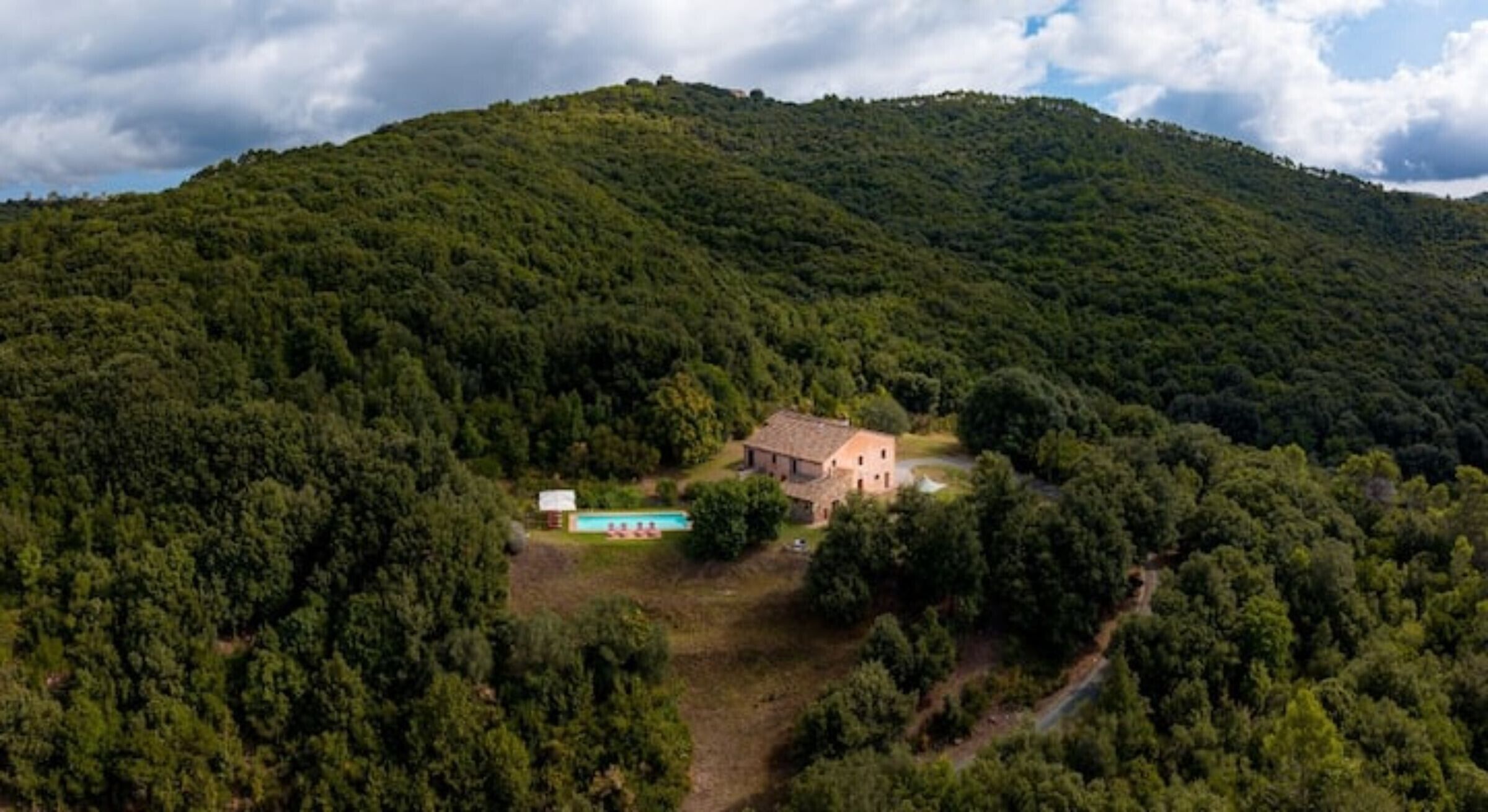 A house surrounded by dense forest, a swimming pool, and a dirt path.