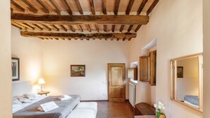 Quarto com teto de madeira, cama com travesseiros e cobertor, mesa de cabeceira com abajur, porta de madeira e espelho na parede.