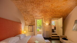 Un dormitorio con techo de ladrillo, una cama con ropa de cama blanca, una mesita de noche con una lámpara y vistas a un baño a través de una puerta abierta.