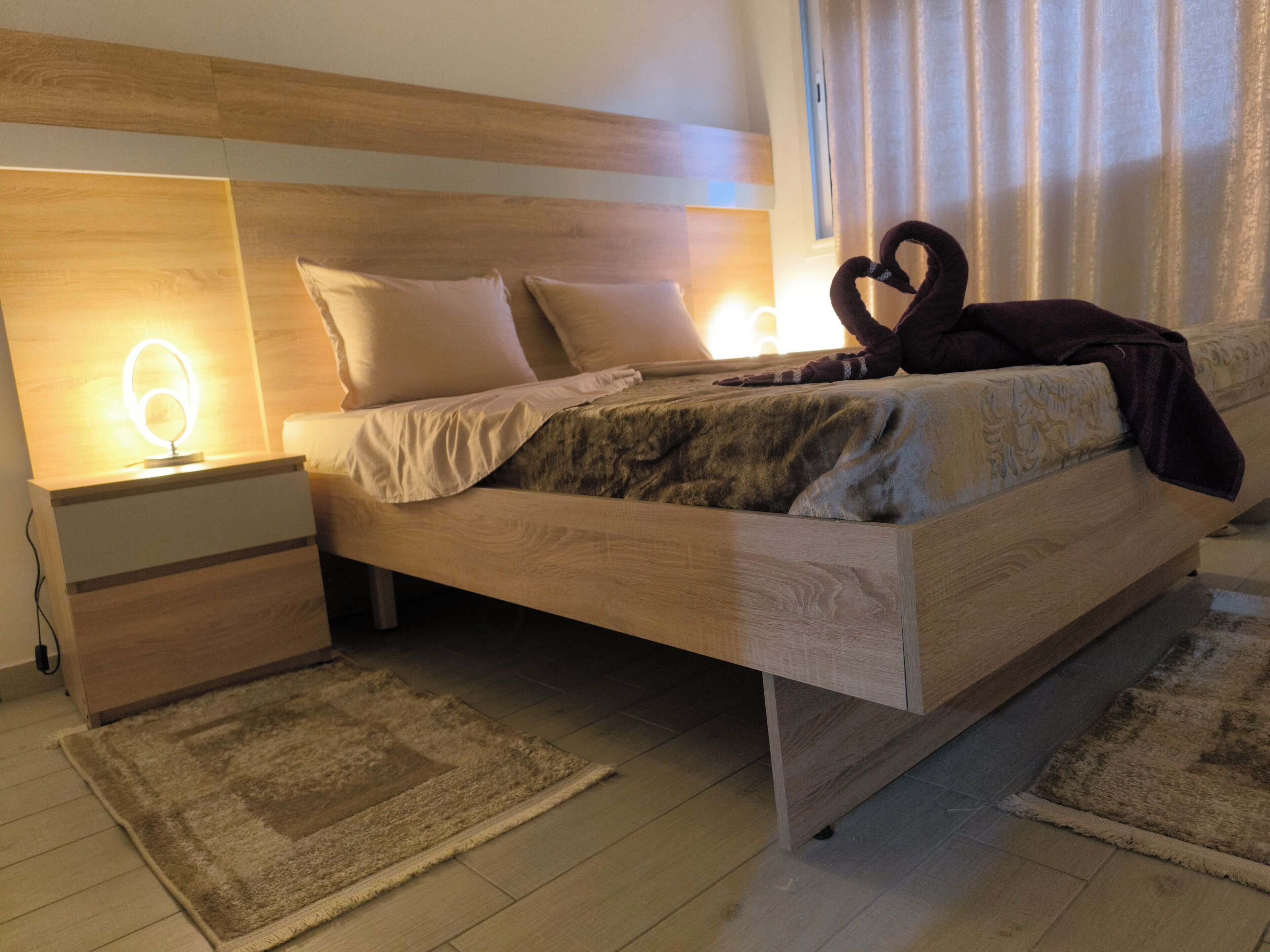 Een moderne slaapkamer met een houten hoofdbord, een bed met een deken en een hartvormig kussen, een nachtkastje met een lamp, en een vloerkleed.