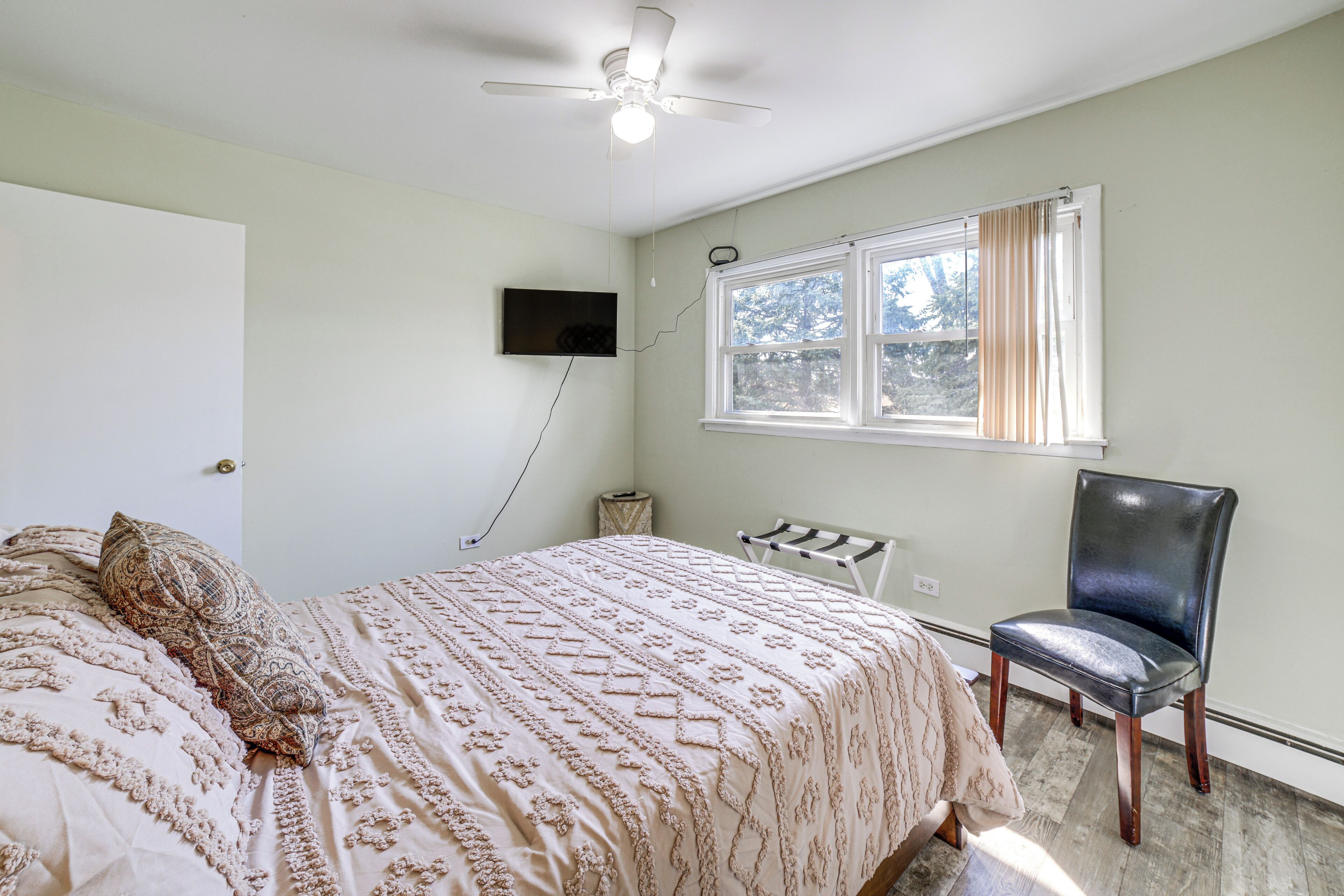 A bedroom with a bed, a chair, a ceiling fan, a window with curtains, and a wall-mounted TV.