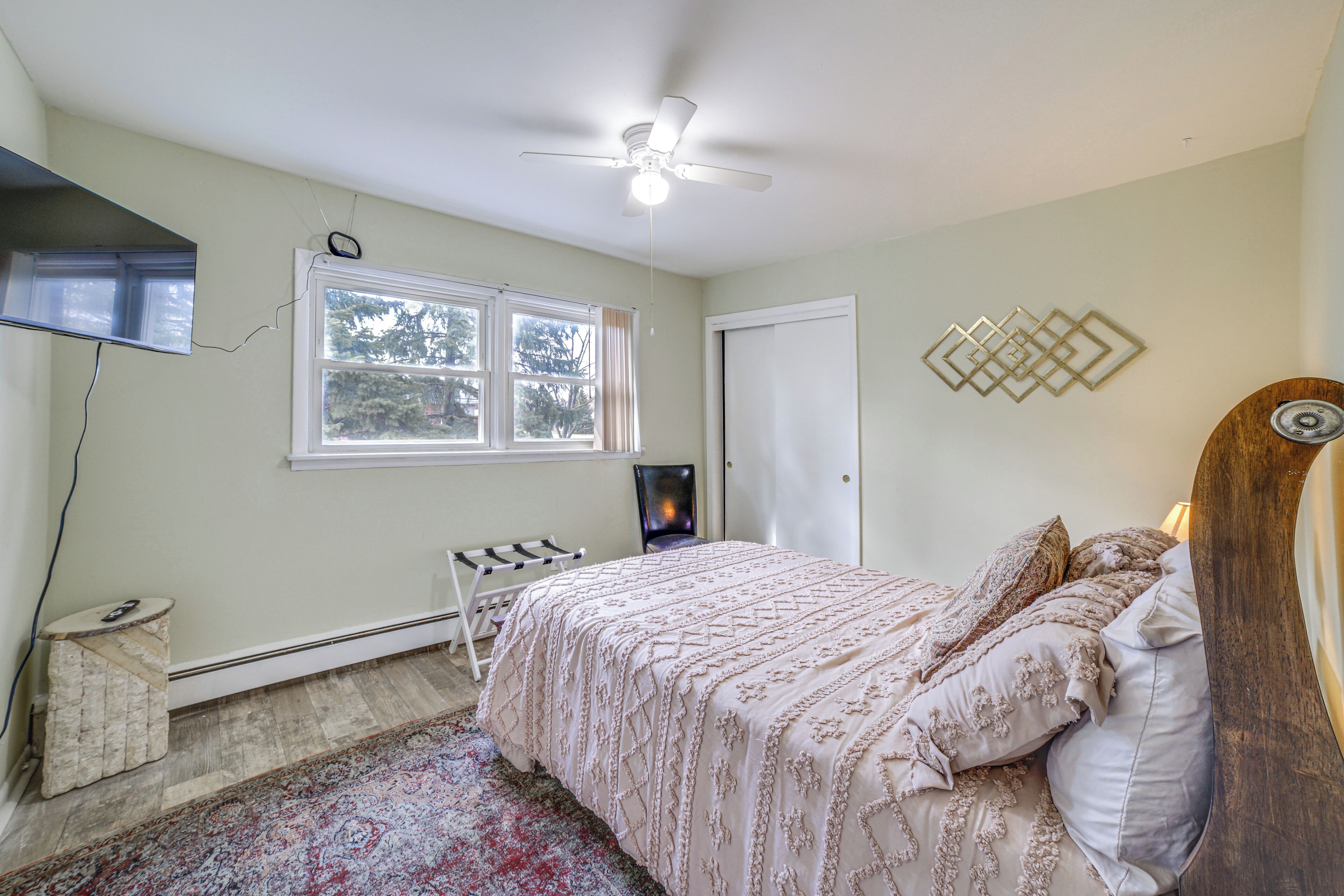 A bedroom with a bed, a ceiling fan, a TV, a window, a radiator, and a wall decoration.