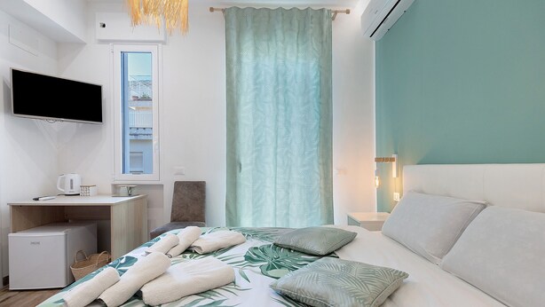 Habitación de hotel con cama, televisor de pantalla plana, escritorio con computadora, refrigerador pequeño y ventana con vista a un edificio.