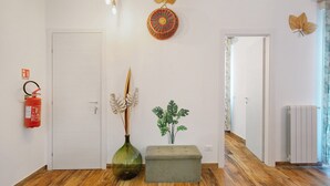 Un pasillo con una puerta blanca, un extintor, un jarrón verde, una planta y un banco gris.