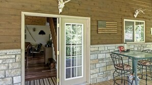A wooden cabin interior with a stone wall, a dining table with chairs, a fireplace, and a door leading to a porch. - Hocking Hills Half Stoned Cabin in the Woods (Nelsonville)