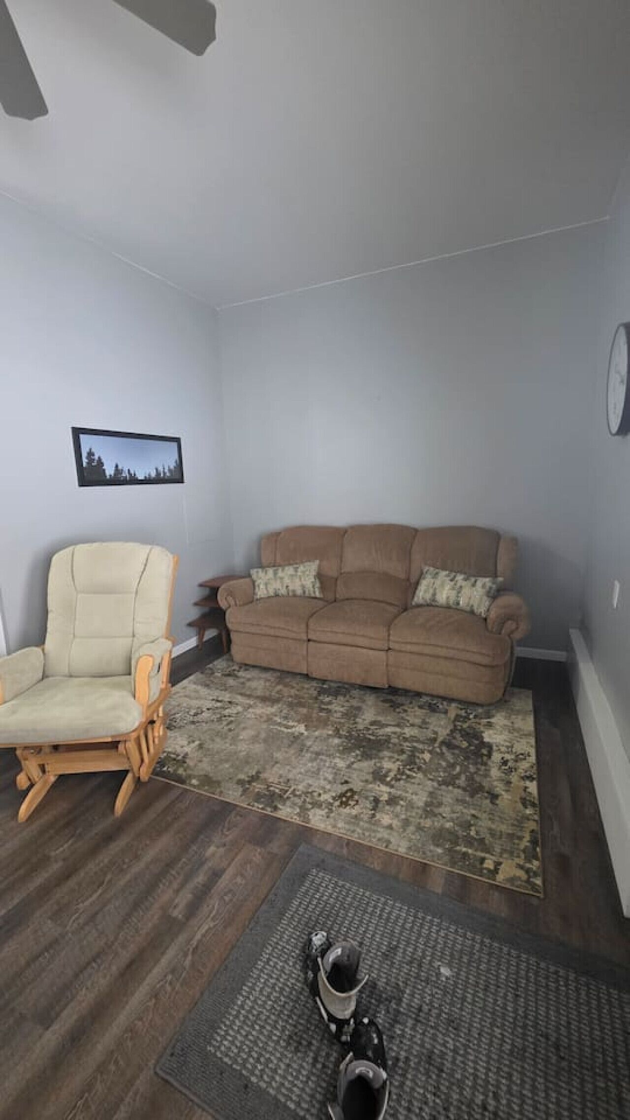 Ein Wohnzimmer mit einem beigen Sessel, einem braunen Sofa mit gemusterten Kissen, einem Teppich und einer Wanduhr.