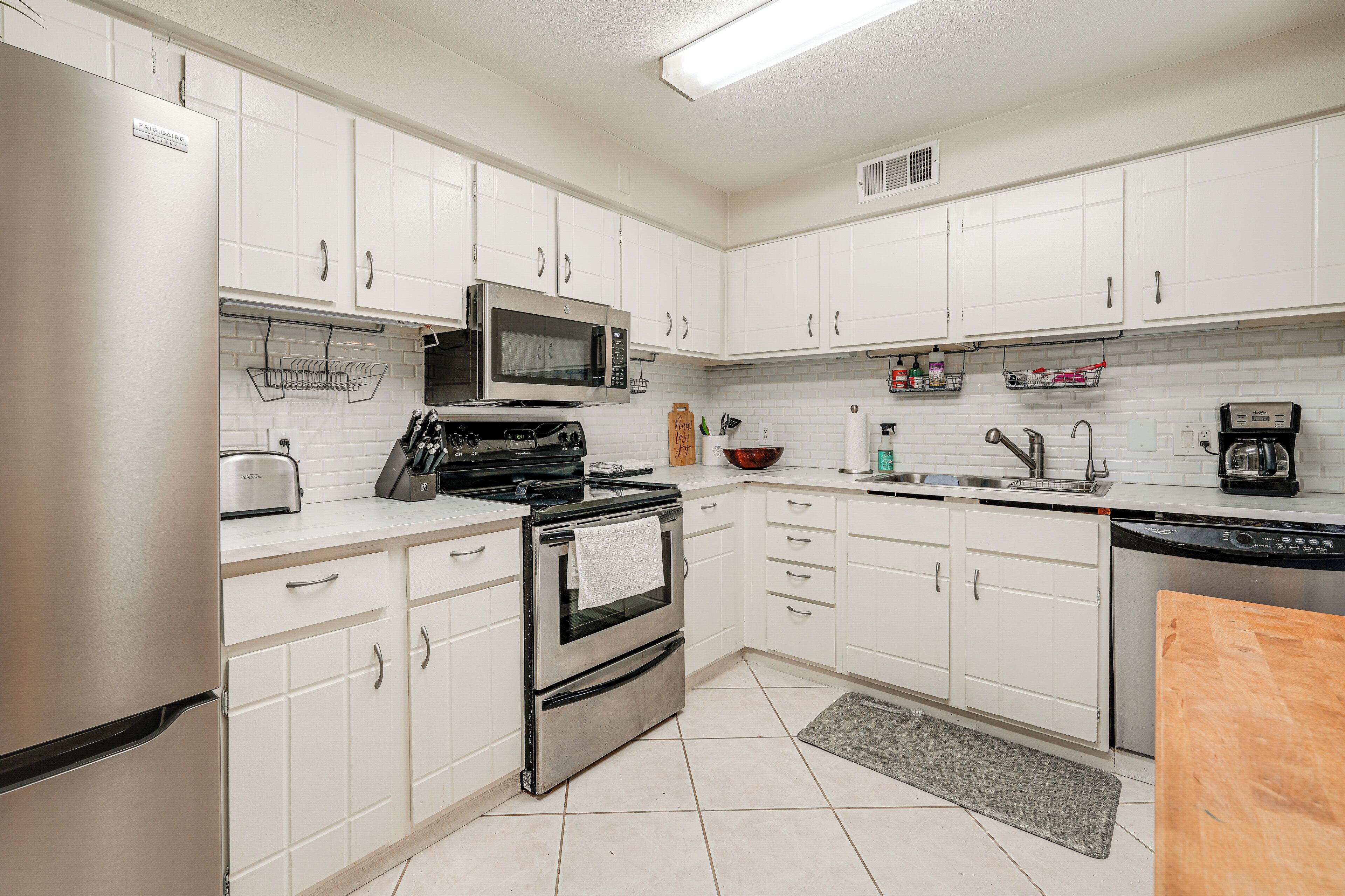 A modern kitchen with white cabinets, a stainless steel refrigerator, a microwave, an oven, a dishwasher, and a wooden cutting board.