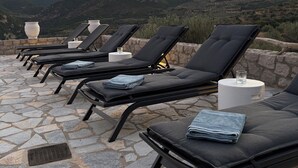 A row of black lounge chairs with towels and glasses on white round tables, set on a stone patio with a mountainous backdrop. - Villa ELIA HOUSE near Nafplio sea and mountain view (Nauplie)