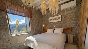A bedroom with a stone wall, a bed with a white bedspread, a nightstand, a wall-mounted lamp, and a window with a view of mountains. - Villa ELIA HOUSE near Nafplio sea and mountain view (Nauplie)