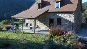 Ein Steinhäuschen mit geneigtem Dach, einer überdachten Terrasse mit Sitzgelegenheiten und einem Garten mit verschiedenen Pflanzen.
