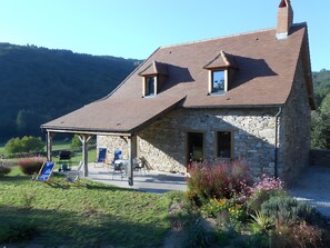 Une maison en pierre avec un toit en pente, une terrasse couverte avec des chaises et un jardin où poussent différentes espèces de plantes.