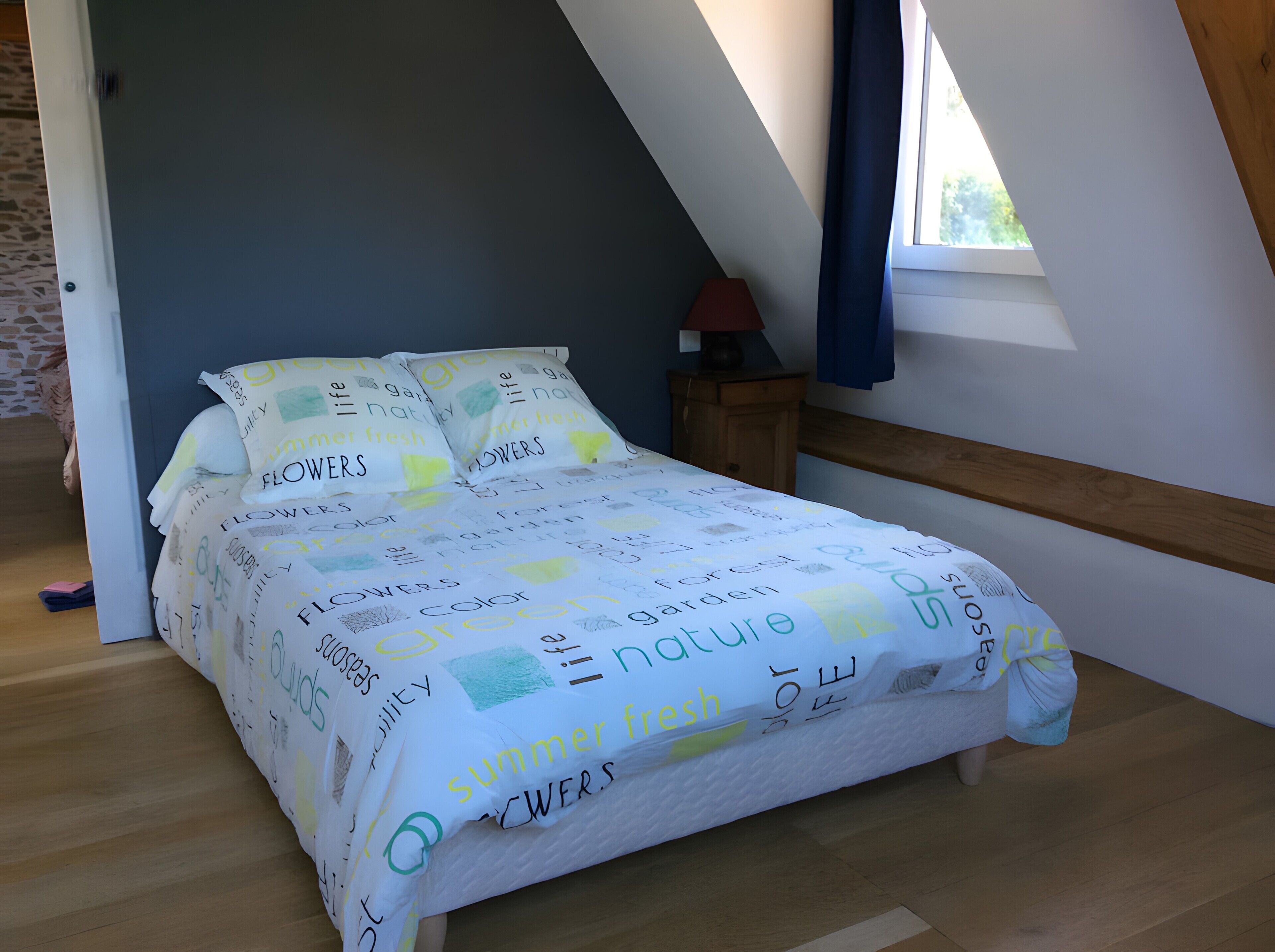 Une chambre avec un plafond en pente, un lit recouvert d’une couvre-lit à motifs floraux, une table de chevet avec une lampe et une fenêtre avec des rideaux.