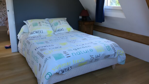 Ein Schlafzimmer mit schrägen Wänden, einem Bett mit blumigem Überwurf, einem Nachttisch mit Lampe und einem Fenster mit Vorhängen.