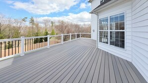 A wooden deck with a white railing, a large window, and a view of trees and sky. - Spacious 4 bd Modern Home with Private Yard & Games (Woodbridge)