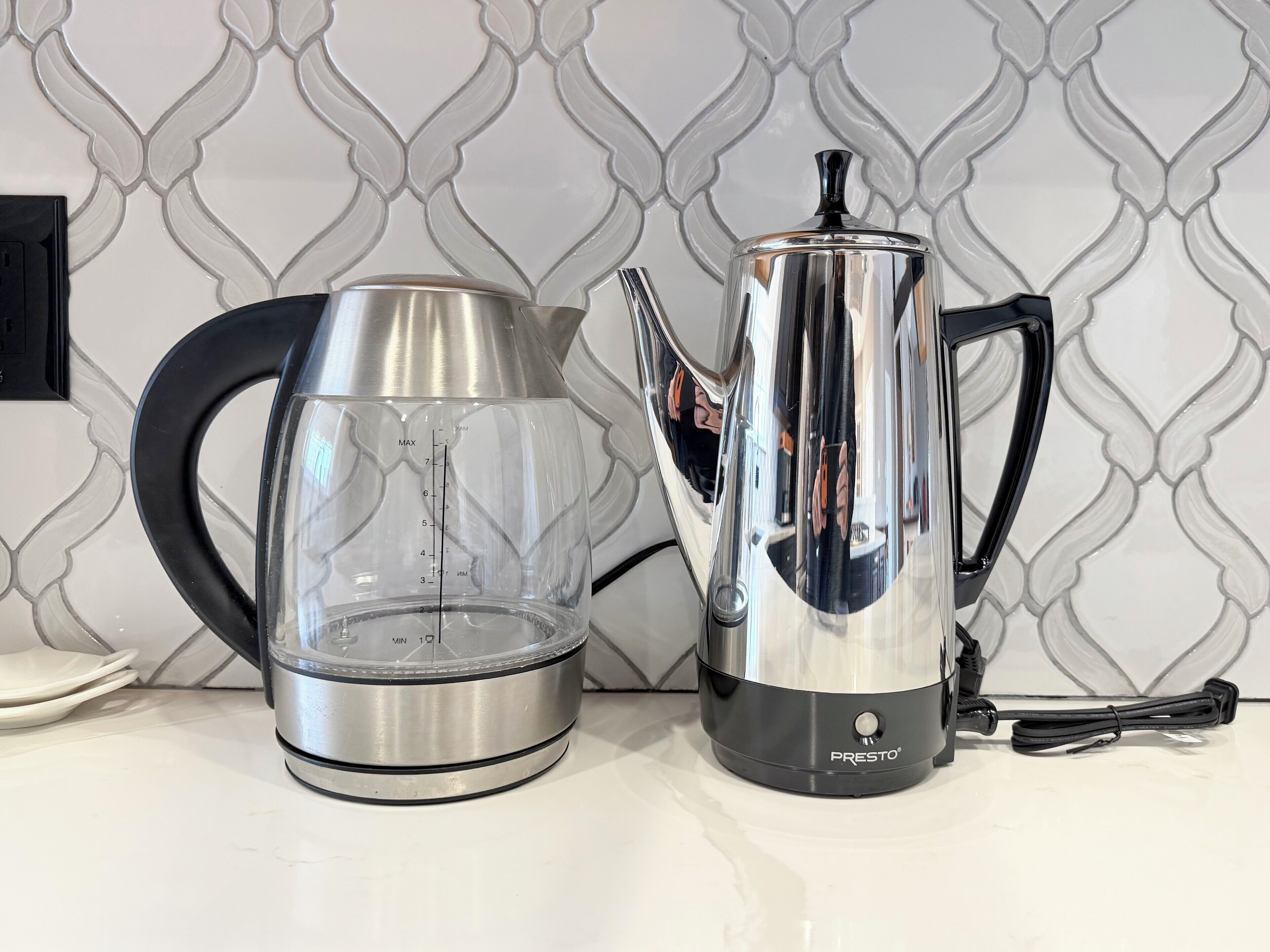 Une bouilloire en acier inoxydable et une carafe en verre sont posées sur un plan de travail, avec un pare-éclaboussures carrelé.