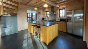 A kitchen with wooden beams, a central island with stools, stainless steel appliances, and a view through the window. - Villa With Sauna & Tub (Den Ham)