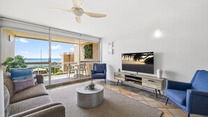 A living room with a TV, two chairs, a coffee table, and a balcony with a view of the ocean. - Look at the beach, Alexandra Headland, sleeps 5 (Alexandra Headland)