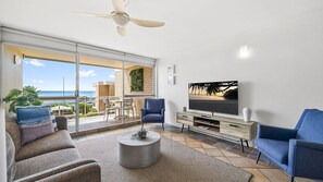 Ein Wohnzimmer mit Fernseher, zwei Sesseln, einem Couchtisch und einem Balkon mit Meerblick.