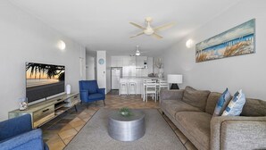 A modern living room with a TV, blue armchair, grey sofa, and a kitchen in the background. - Look at the beach, Alexandra Headland, sleeps 5 (Alexandra Headland)