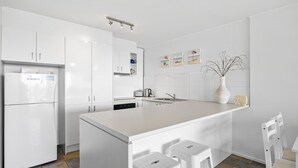 A modern kitchen with white cabinetry, a central island, and a small dining area. - Look at the beach, Alexandra Headland, sleeps 5 (Alexandra Headland)