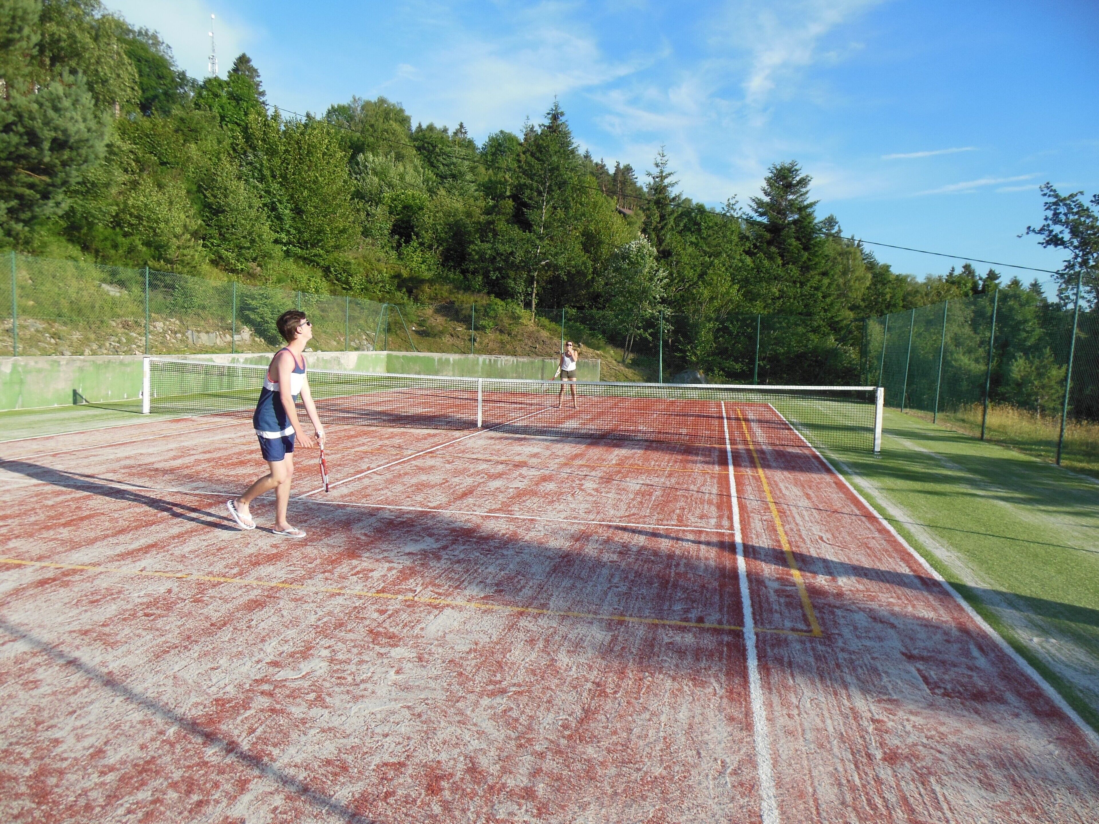 An outdoor tennis court with a net, surrounded by trees and a fence.