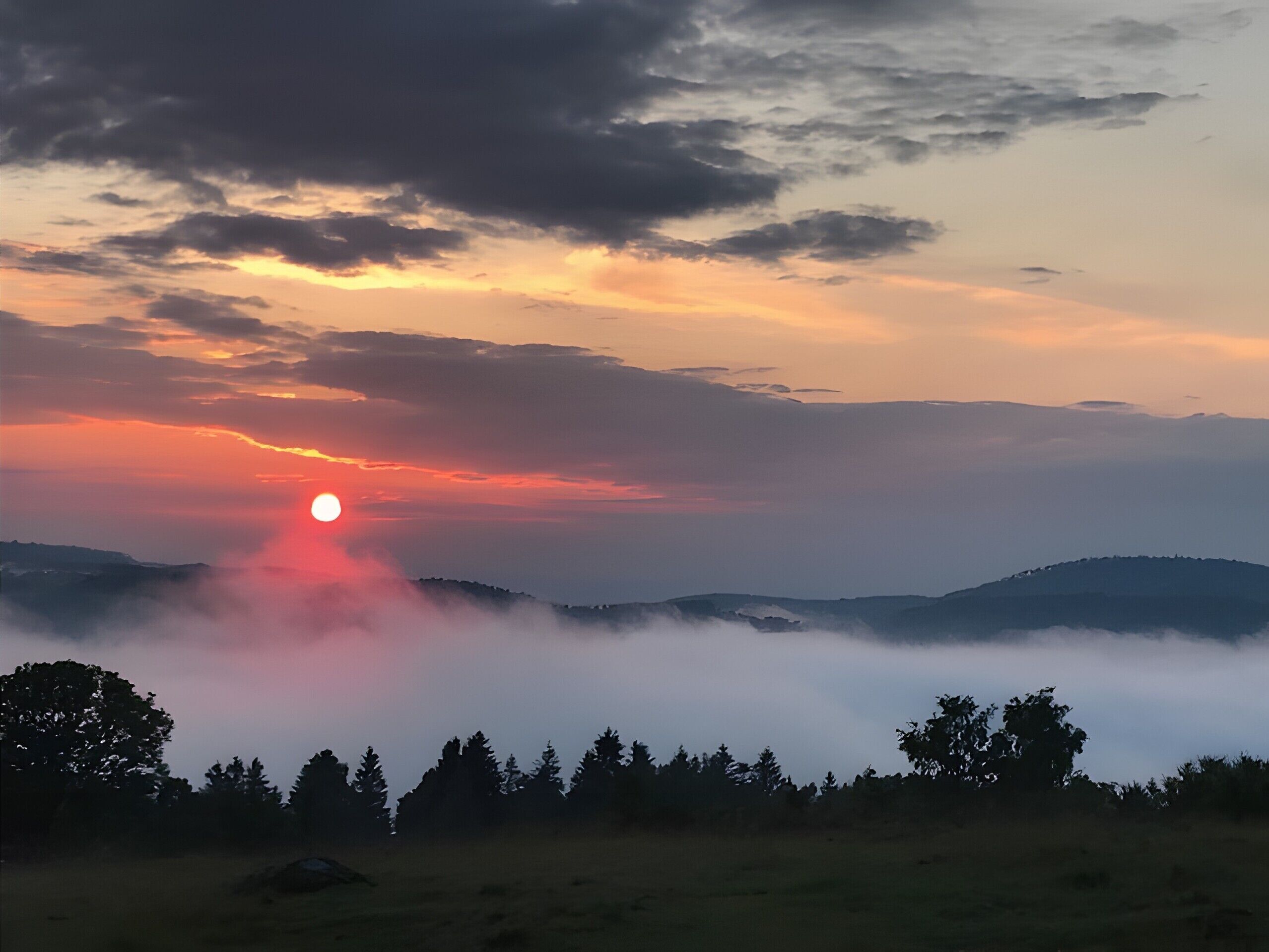 Paisagem com o sol se pondo atrás de colinas, rodeada por nuvens e árvores.