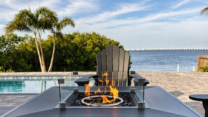 Ein Tisch am Pool mit integriertem Feuerbereich, ein Stuhl und Blick auf das Meer und Palmen.