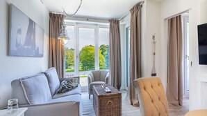 A modern living room with a sofa, armchair, and a small table, large windows with curtains, and a flat-screen TV. - 4 Star Holiday Home in Travemunde (Lübeck)