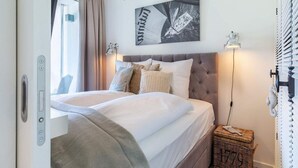 A bedroom with a bed, a headboard, a wall-mounted artwork, a bedside lamp, a window with curtains, and a wicker basket. - 4 Star Holiday Home in Travemunde (Lübeck)