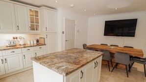 Una cocina con armarios blancos, encimera de granito, una mesa de comedor con sillas y un televisor de pantalla plana montado en la pared.