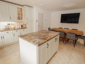 A kitchen with white cabinets, a granite countertop, a dining table with chairs, and a flat-screen TV mounted on the wall. - Willows Lodge at Carreg Llwyd (Cardiff)