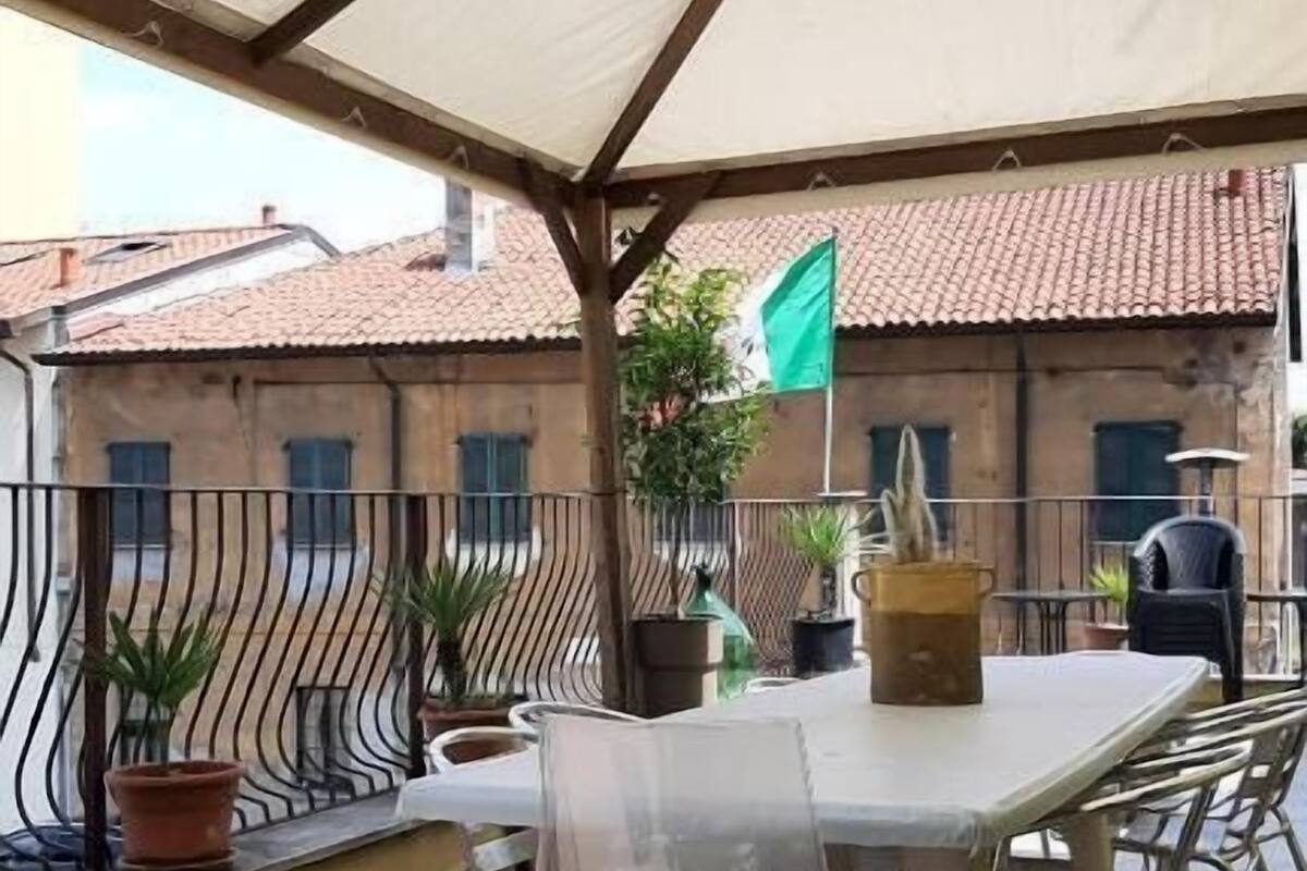 Une terrasse extérieure couverte, avec une table et des chaises, des plantes en pot et une vue sur les bâtiments en arrière-plan.