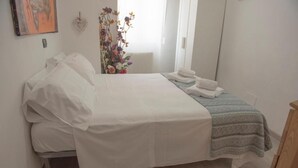 Un letto rifatto con lenzuola bianche, un piccolo vaso con fiori e una finestra che lascia entrare luce naturale.