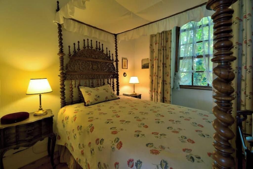 A bedroom with a four-poster bed, two bedside lamps, a floral bedspread, and a window with curtains.
