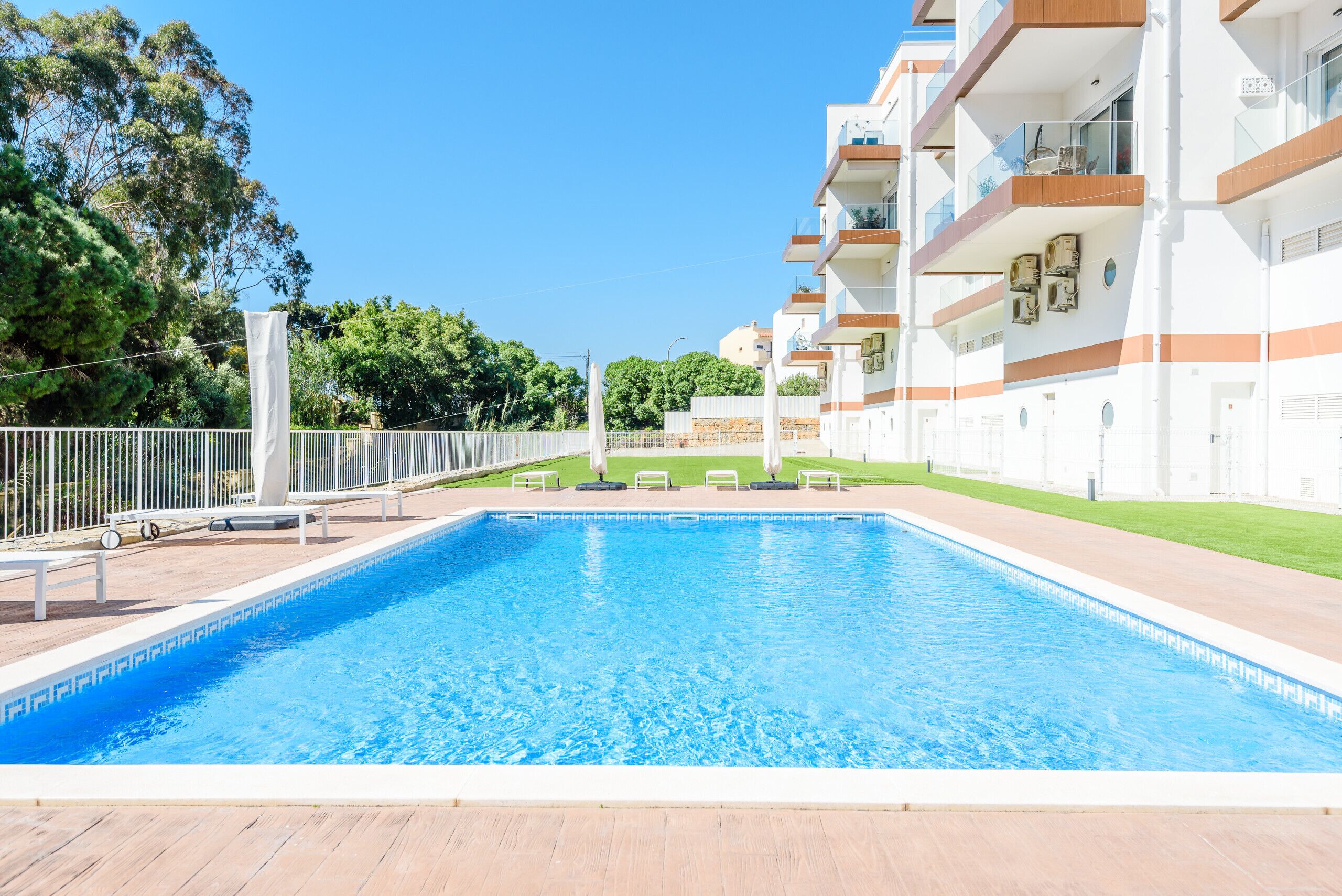Une piscine dotée d’une terrasse en bois, entourée d’une clôture blanche et jouxtant un immeuble à plusieurs étages avec des balcons.